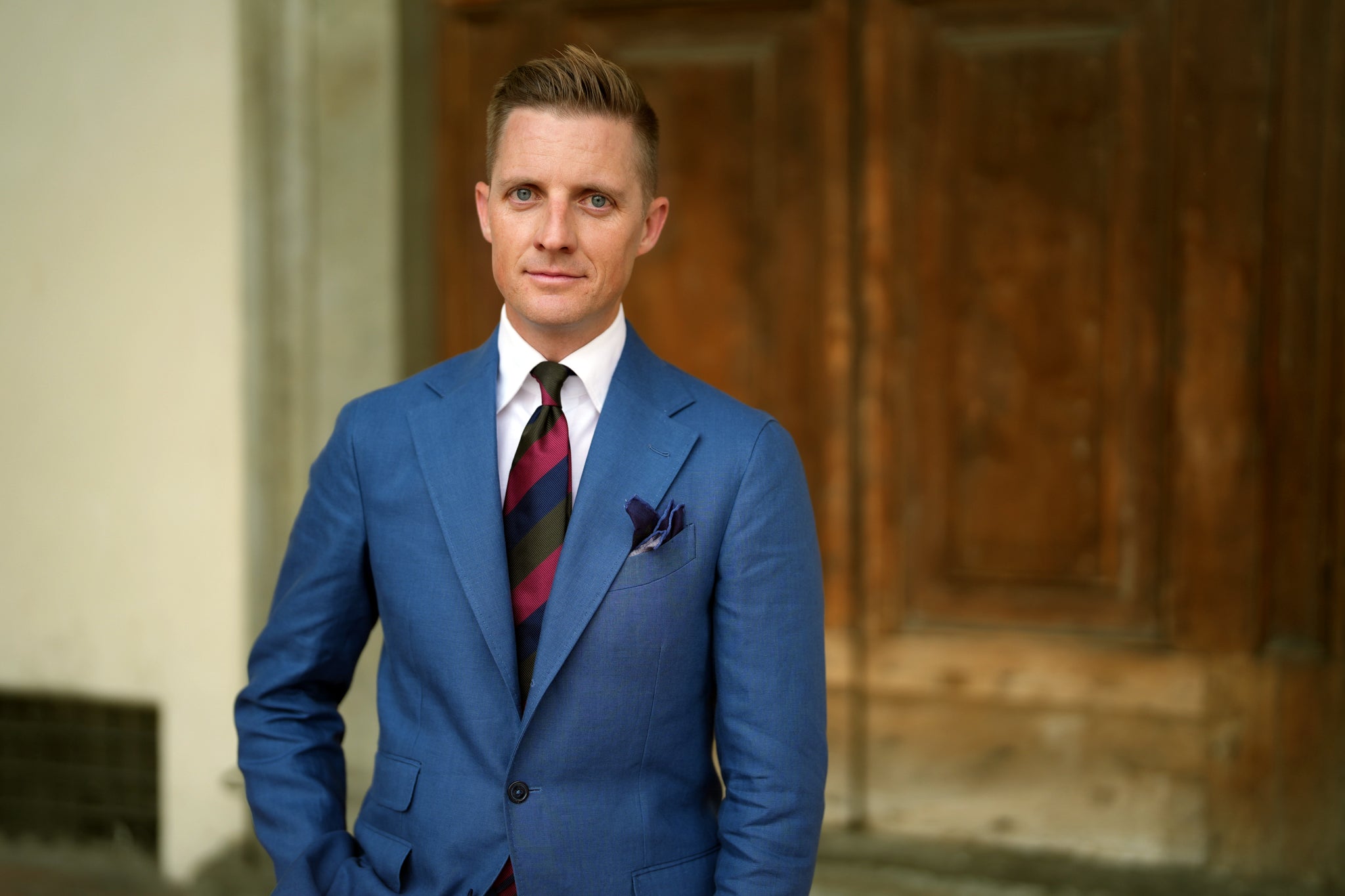 Man wearing a Beckett & Robb blue single-breasted suit with notch lapels, styled with a white shirt, bold striped tie, and purple pocket square, standing in front of a wooden doorway.