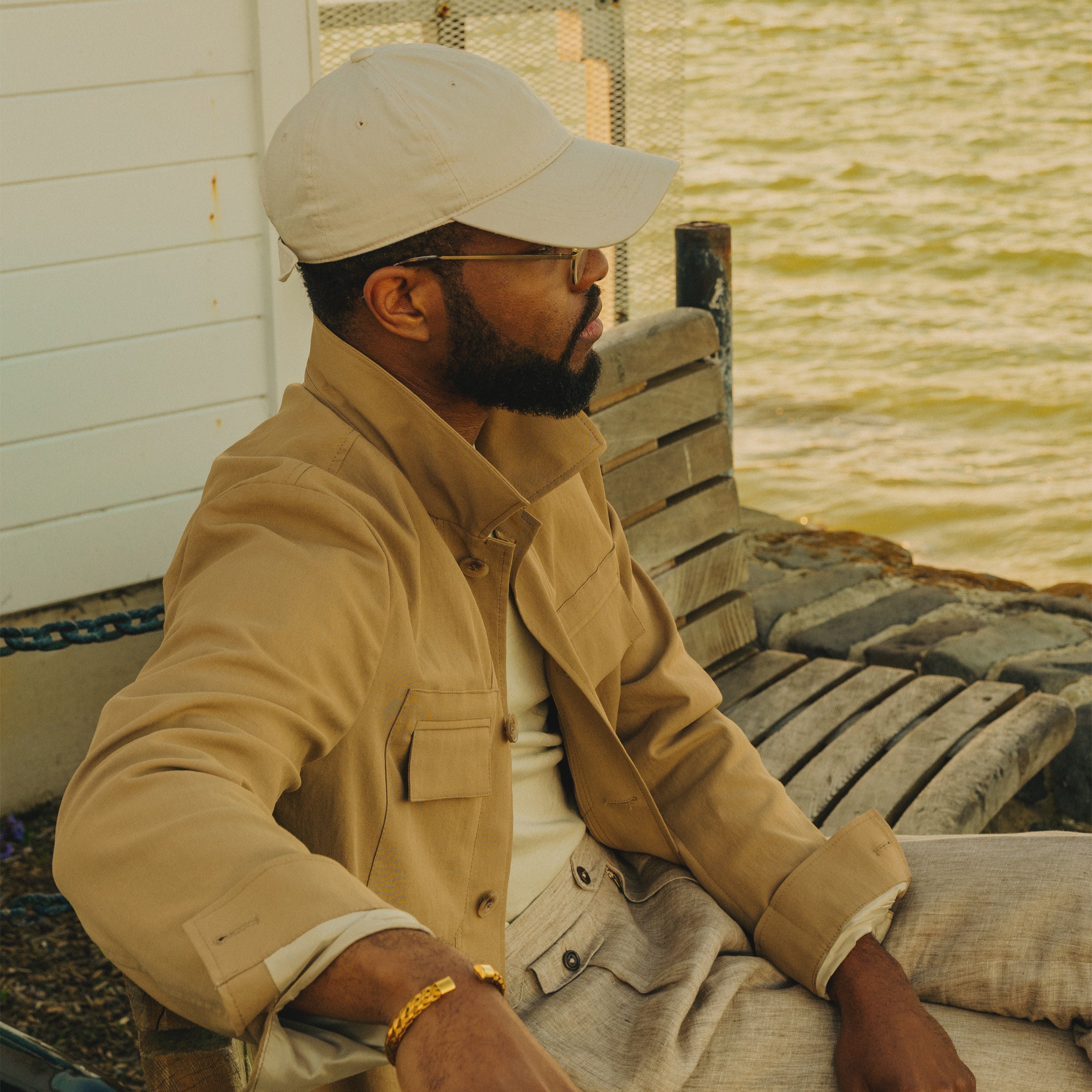 Man seated on a wooden bench by the water, wearing a tan WJ-001 work jacket, cream T-shirt, beige trousers, and a light baseball cap