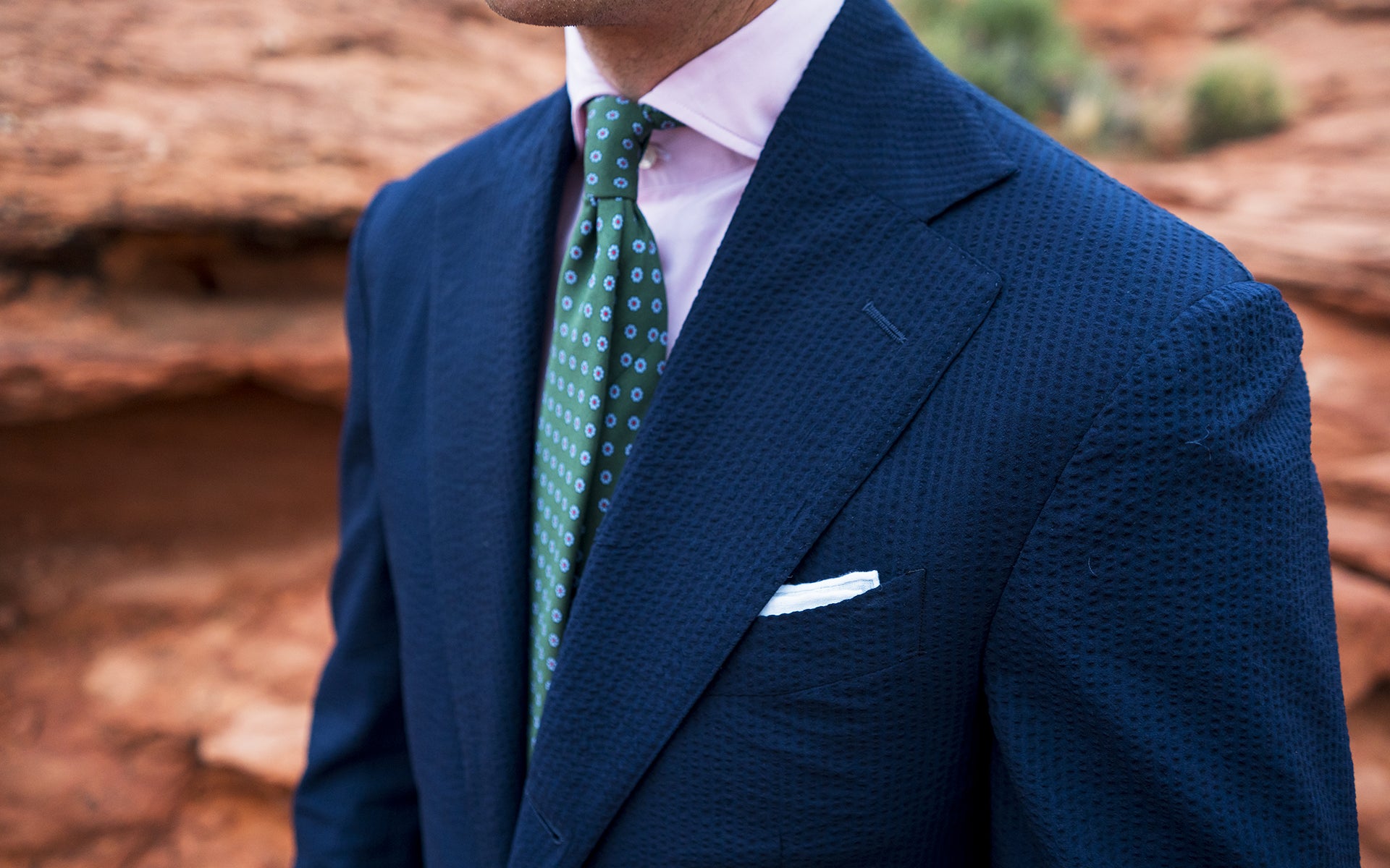 close-up of a man wearing a navy seersucker suit with a white pocket square, green patterned tie, and pale pink dress shirt, set against a red rock background.